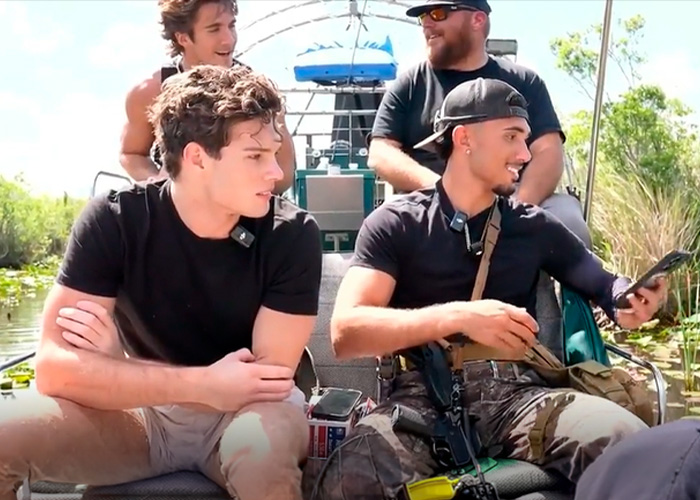 Four men outdoors on a boat, two wearing black shirts, one checking a phone, related to looksmaxxing influencer arrest.