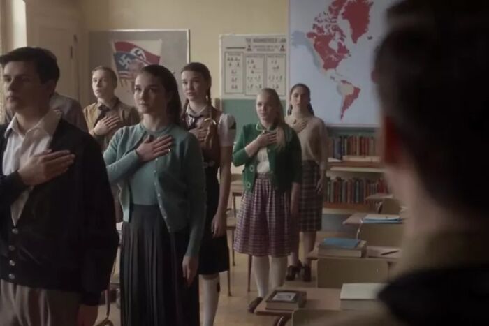 Students in a vintage American classroom standing with hands over hearts during the Pledge of Allegiance ceremony.