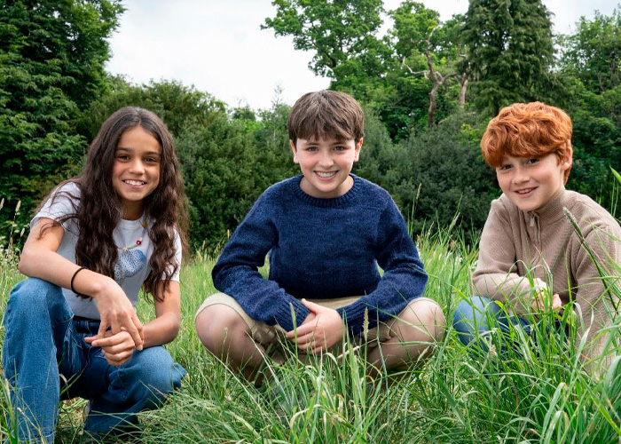 Three kids smiling and crouching in a grassy field with trees in the background, drone over Harry Potter set concept.