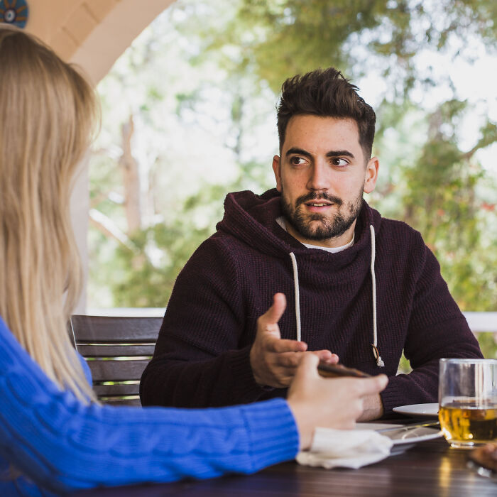 Man wearing a maroon hoodie gesturing while mansplaining during a conversation with a woman at an outdoor table.