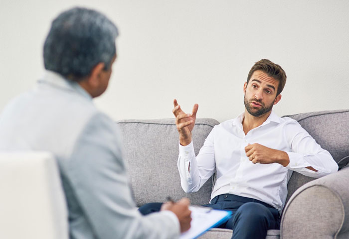 Man in white shirt explaining work-life balance to colleague in suit during a serious office conversation on a gray couch.