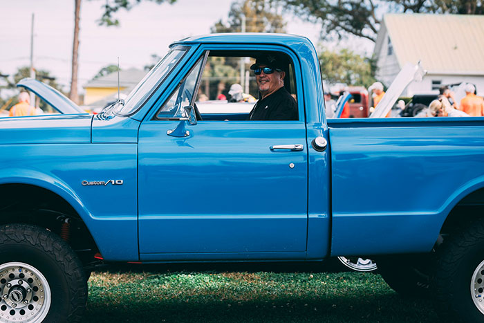 Man wearing sunglasses sitting in a blue vintage truck, with people and trees blurred in the background, eerie unexplained scene.