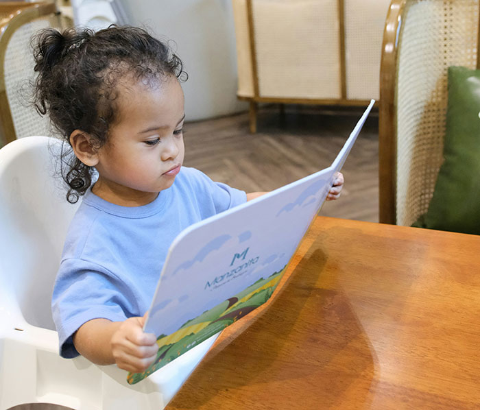 Young child sitting at a table reading a colorful book, capturing a quiet moment of thought-provoking curiosity.