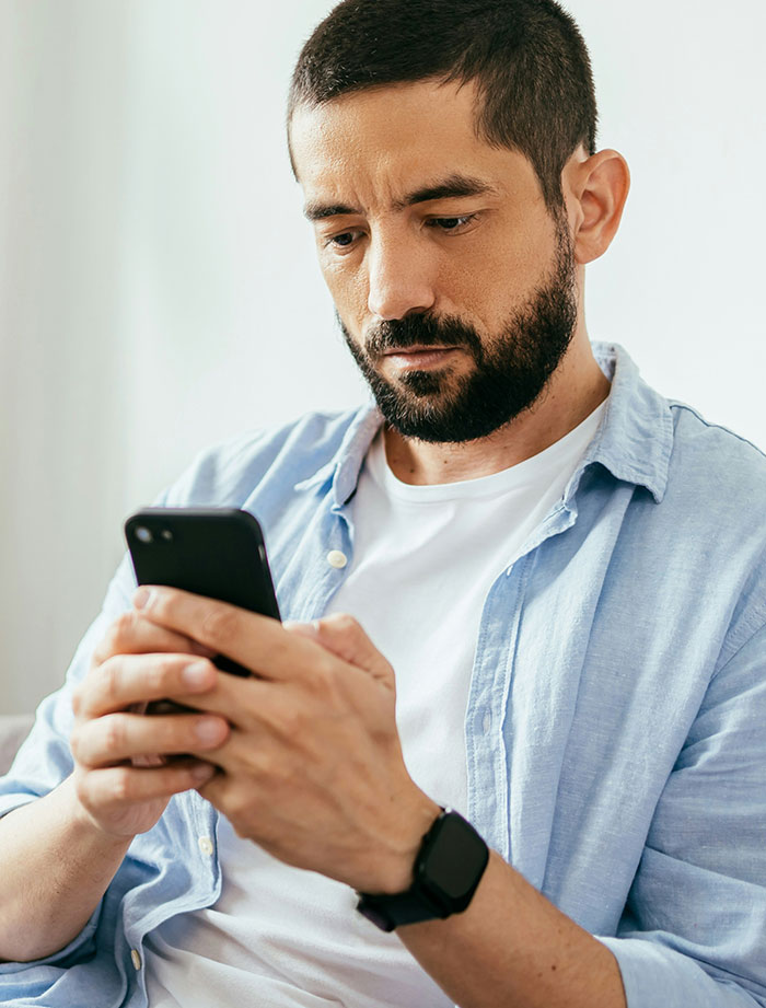Man with a beard wearing a light blue shirt, focused on his phone while reading eerie unexplained stories.