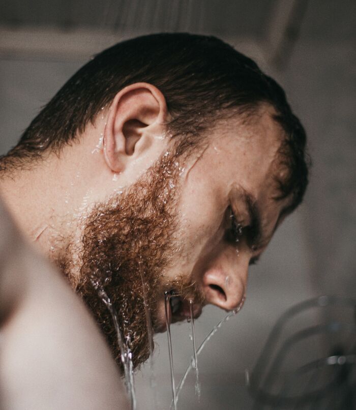 Man with beard and wet hair leaning forward with water streaming down his face in a close-up profile shot.