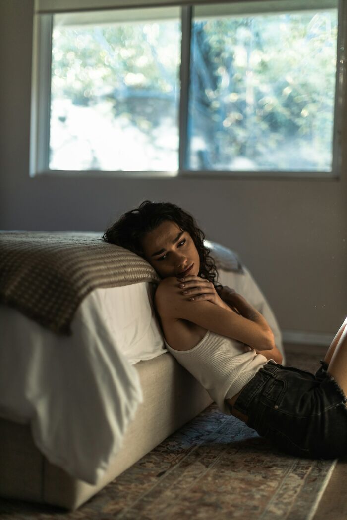 Young woman sitting on the floor leaning against a bed in a dim room, reflecting on American things believed only in movies.