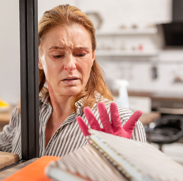 Woman in striped shirt wearing pink gloves, looking concerned while cleaning a dusty air filter in a home environment.