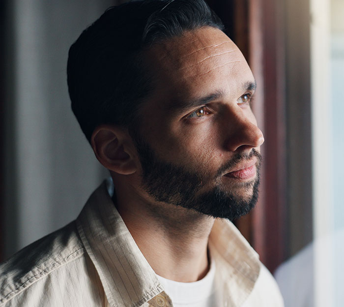 Man with beard and dark hair reflecting deeply by a window, symbolizing moments men drop the red pill mindset.
