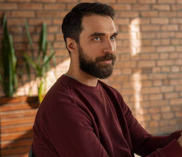 Bearded man in a maroon sweater thoughtfully reflecting, representing men who drop the red pill mindset in a calm indoor setting.