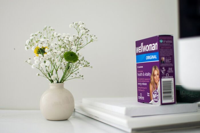 Box of Wellwoman health supplements on white books next to vase with flowers, representing shaky industries facade concept.