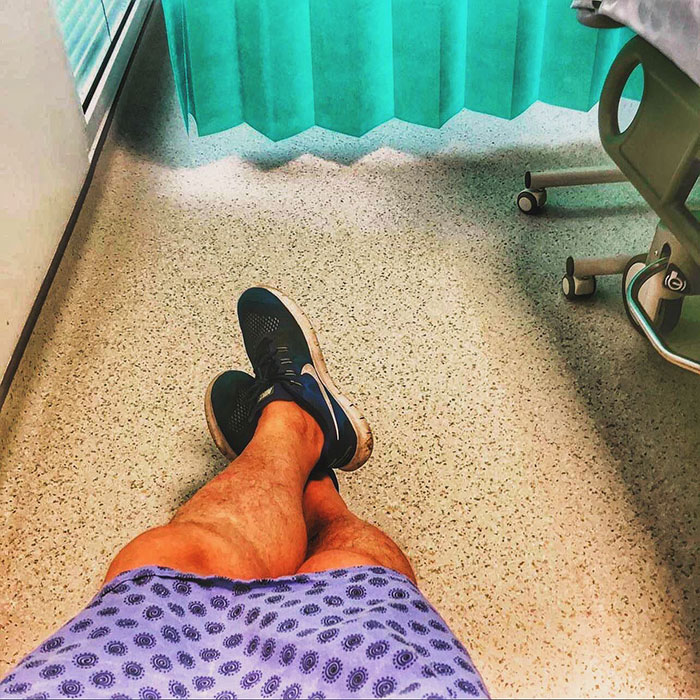 Person wearing patterned hospital gown and black sneakers sitting with legs crossed on speckled floor near medical curtain and chair wheels.