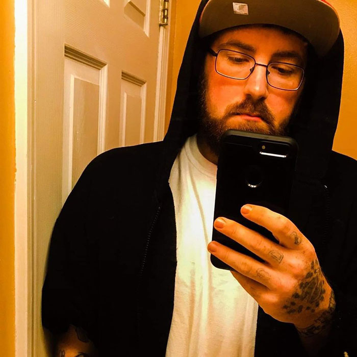 Man with beard and glasses wearing a black hoodie and cap, taking a mirror selfie on a smartphone, owner of tiniest package concept.