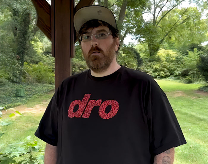 Man wearing black dro t-shirt and beige cap standing outdoors under a wooden structure, representing owner of tiniest package.