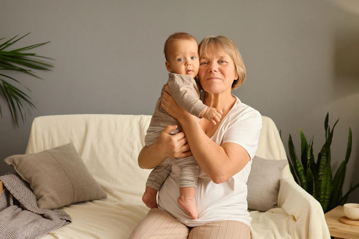 Older woman holding baby on couch in living room, illustrating anxiety linked to new mom and in-laws visits.