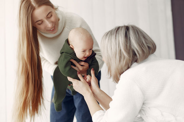 New mom holding baby while interacting with mother-in-law, depicting anxiety and family tension during visits.