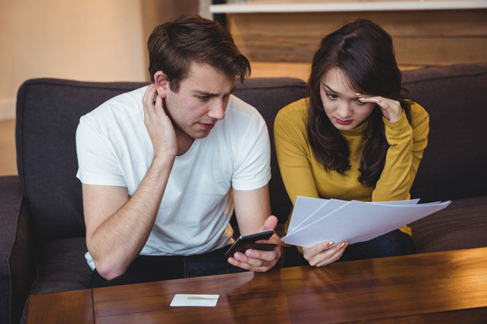 Young couple sitting on a couch, reviewing bills and phone, worried about debt and considering bankruptcy options.