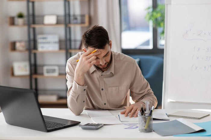 Man sitting at desk looking stressed while calculating debt with papers and laptop representing mil wants file bankruptcy debt concept