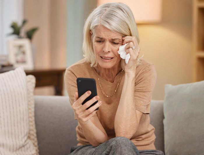 Older woman wiping tears while looking at phone, appearing worried about bankruptcy and debt issues at home.