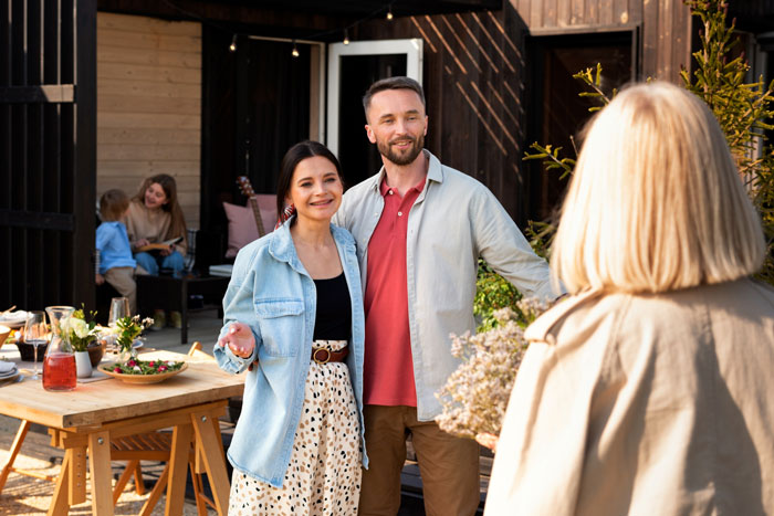 Couple welcoming a friend at a backyard gathering, with outdoor dining table and children in the background.