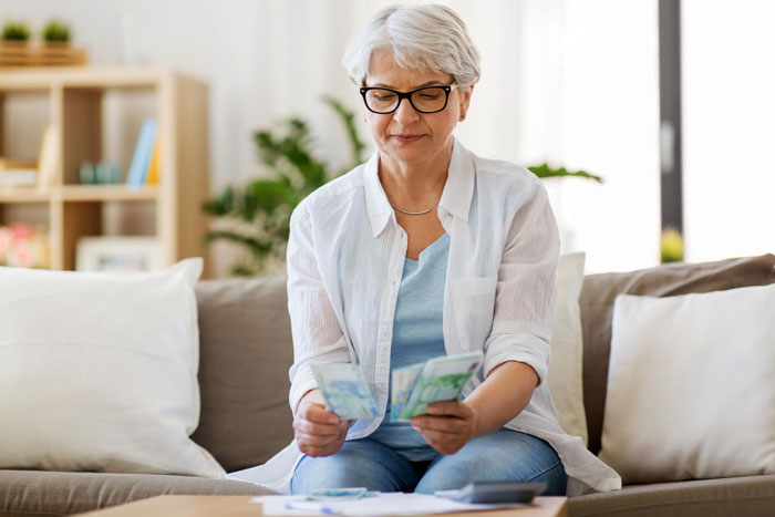 Older woman sitting on a couch counting money with a concerned expression related to bankruptcy debt issues at home.