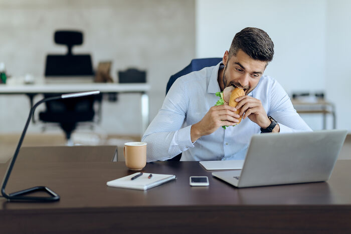 Man eating a sandwich at his desk while working on a laptop, embodying like this generation's moon landing moment