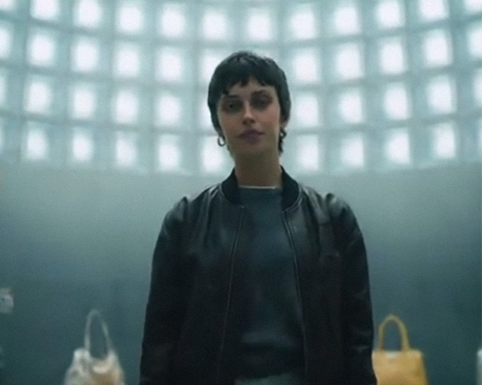 Model standing in a dimly lit room wearing a black jacket, linked to fashion brand plagiarism and AI photo controversy.