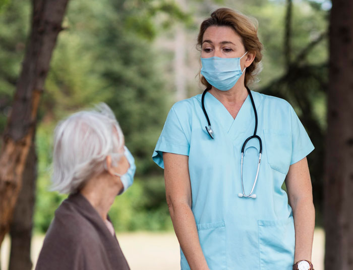 Healthcare worker wearing mask talks to elderly woman outdoors, highlighting mom work and family relationship support.