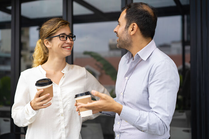 Two colleagues holding coffee cups, smiling and chatting outside an office, showing friendly but potentially nasty behavior.