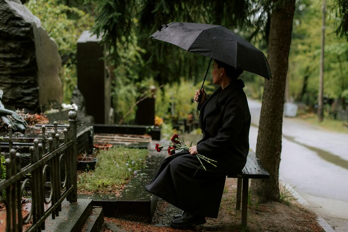 Person sitting on bench at rainy cemetery holding umbrella and flowers, reflecting on the nasty side of human nature.