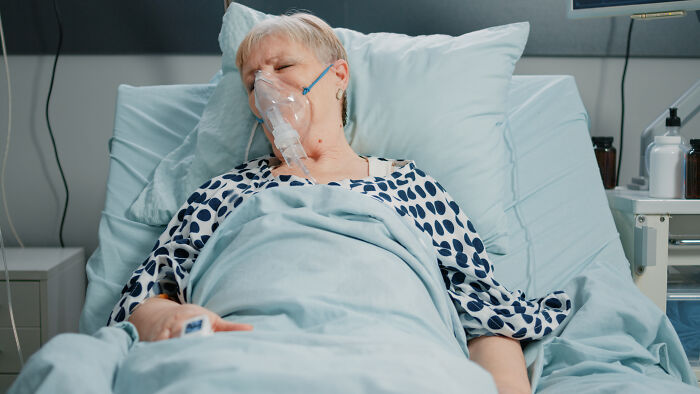 Elderly woman in hospital bed wearing an oxygen mask, looking weak and resting with blue bedding.