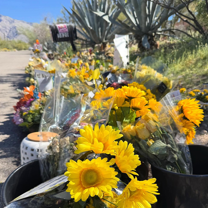 Bright yellow flowers placed along a roadside memorial in the Nancy Guthrie case with gloves nearby.