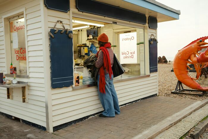 Person wearing a red hat and scarf at a seaside kiosk near large shrimp statues on a cloudy day.