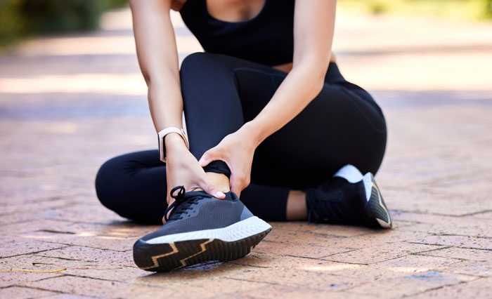 Woman in black athletic wear sitting outdoors, holding ankle in discomfort after possible injury or strain