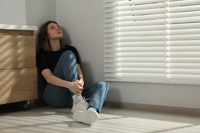 Young woman sitting on floor by window blinds, looking unsettled and deep in thought about helpful neighbor caretaker lies.