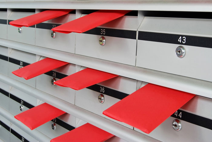 Red wedding invites sticking out of multiple silver mailboxes in an apartment building with numbered slots.