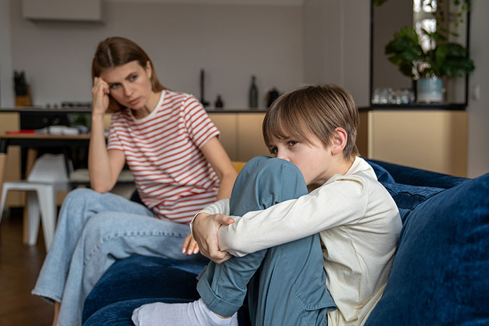 11-year-old child sitting on couch looking upset while mom in striped shirt appears frustrated in living room setting