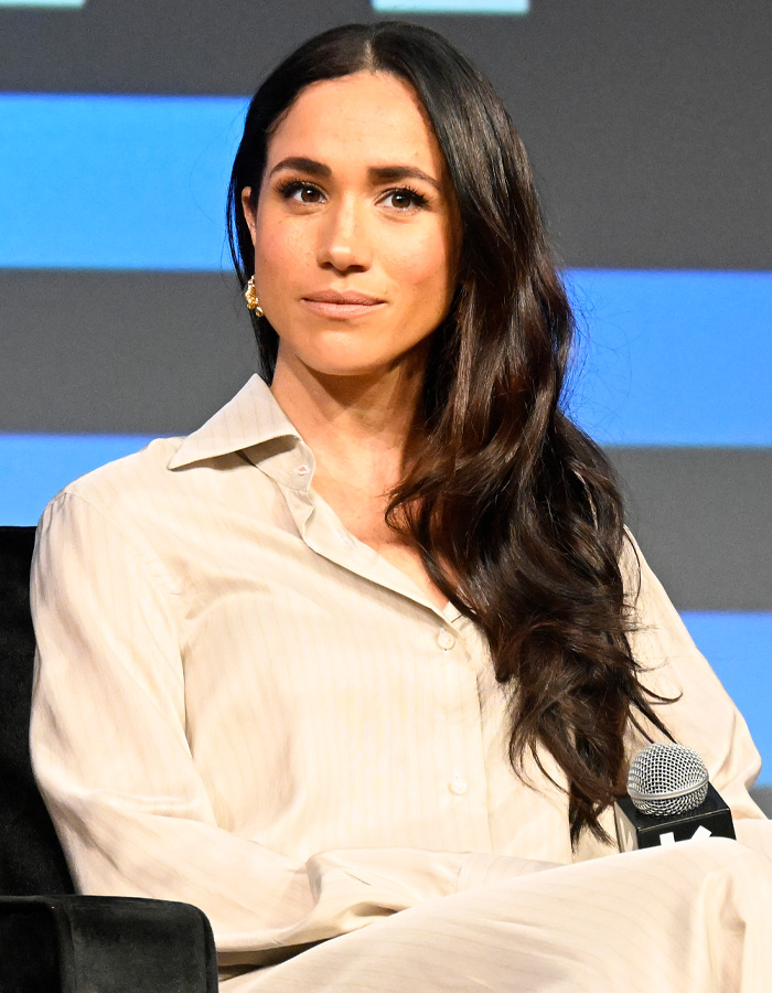 Meghan Markle seated during a panel discussion, wearing a cream blouse and holding a microphone at an event.