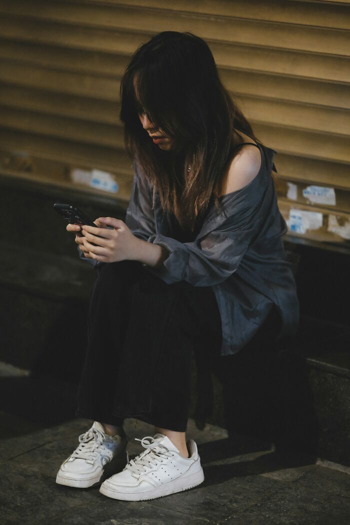 Young woman sitting alone in dim light, focused on her phone, illustrating moments people destroyed their own lives.