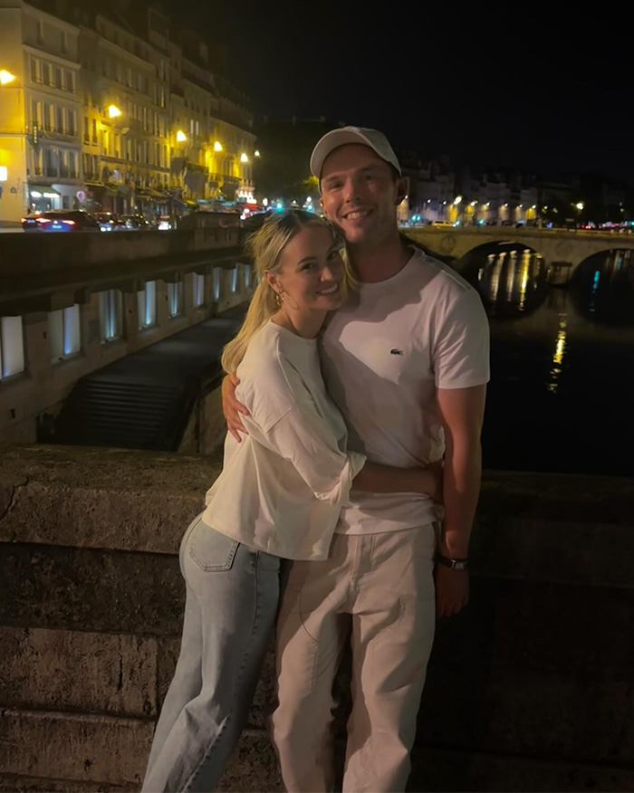 Model Bryana Holly and husband Nicholas Hoult smiling and hugging at a nighttime cityscape by the river.