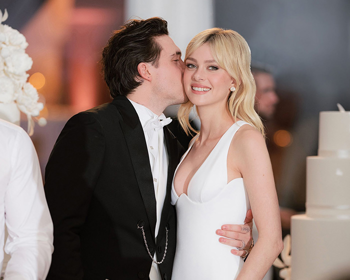 Nicola Peltz smiling in white dress as Brooklyn Beckham kisses her cheek at a formal event with blurred background.