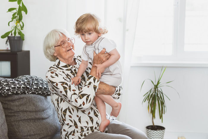 Elderly woman holding toddler in a bright living room, highlighting themes of violent niece injures and enabler family issues.