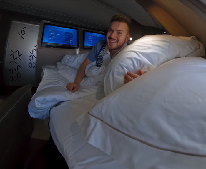 Influencer lying in luxury airplane bed, smiling with screens behind, highlighting refusal to fly economy during conflict.