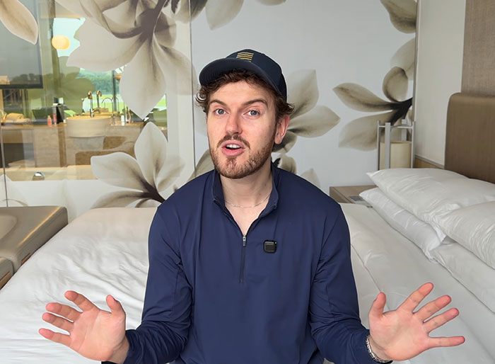 Influencer in a cap and navy top sitting on a bed, expressing frustration about being stranded due to refusing to fly economy.