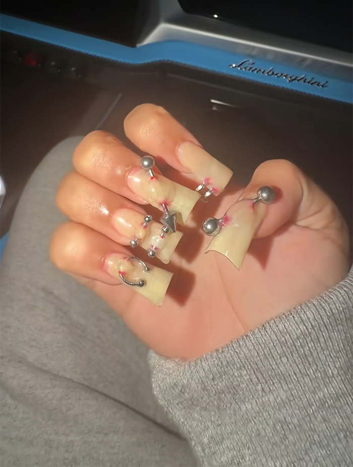 Hand with spiked pierced manicure featuring large metal studs and rings on long n**e-colored nails.