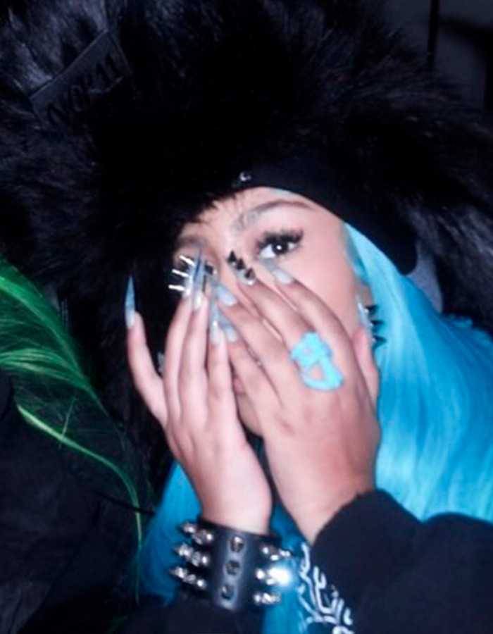 Person with blue hair and spiked, pierced manicure covering part of their face, wearing a black furry hat and studded bracelet.