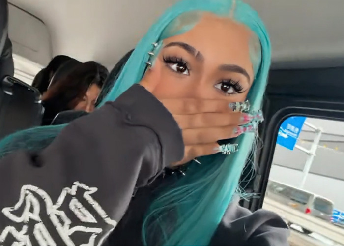 Person with turquoise hair covering mouth, showing spiked pierced manicure inside a vehicle with other passengers visible.