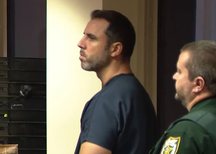 Man in dark blue shirt standing next to a law enforcement officer during a courtroom hearing related to nurse's chilling final text case