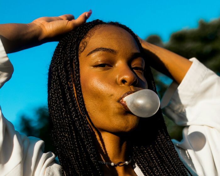 Young woman with braided hair blowing a bubblegum bubble outdoors, capturing a lighthearted culture shock moment.