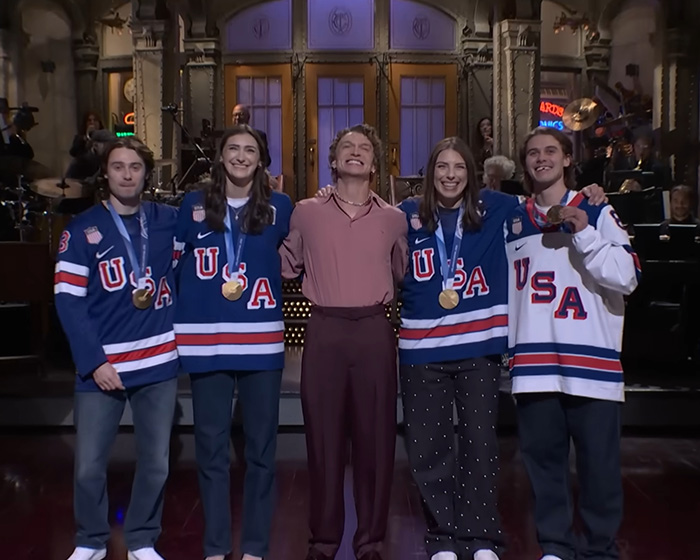 Olympic hero Jack Hughes smiling awkwardly with teammates wearing USA hockey jerseys and gold medals on stage. Olympic hero Jack Hughes smiling awkwardly with teammates wearing USA hockey jerseys and gold medals on stage.
