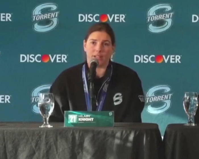 Athlete at a press conference wearing a black jacket with logos, speaking into a microphone during an interview session. Athlete at a press conference wearing a black jacket with logos, speaking into a microphone during an interview session.